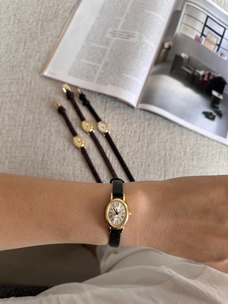 K&ouml;nnte beinhalten: Eine goldfarbene Armbanduhr mit schwarzem Lederarmband wird am Handgelenk getragen. Die Uhr hat ein wei&szlig;es Zifferblatt mit r&ouml;mischen Ziffern. Drei weitere Uhren mit goldenen Zifferbl&auml;ttern und Lederarmb&auml;ndern sind im Hintergrund zu sehen.
