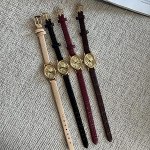 K&ouml;nnte beinhalten: Vier Damenuhren mit goldfarbenen Geh&auml;usen und Lederarmb&auml;ndern in Beige, Schwarz, Burgunderrot und Braun. Jede Uhr hat ein rundes Zifferblatt mit r&ouml;mischen Ziffern und einer kleinen Sekundenscheibe.