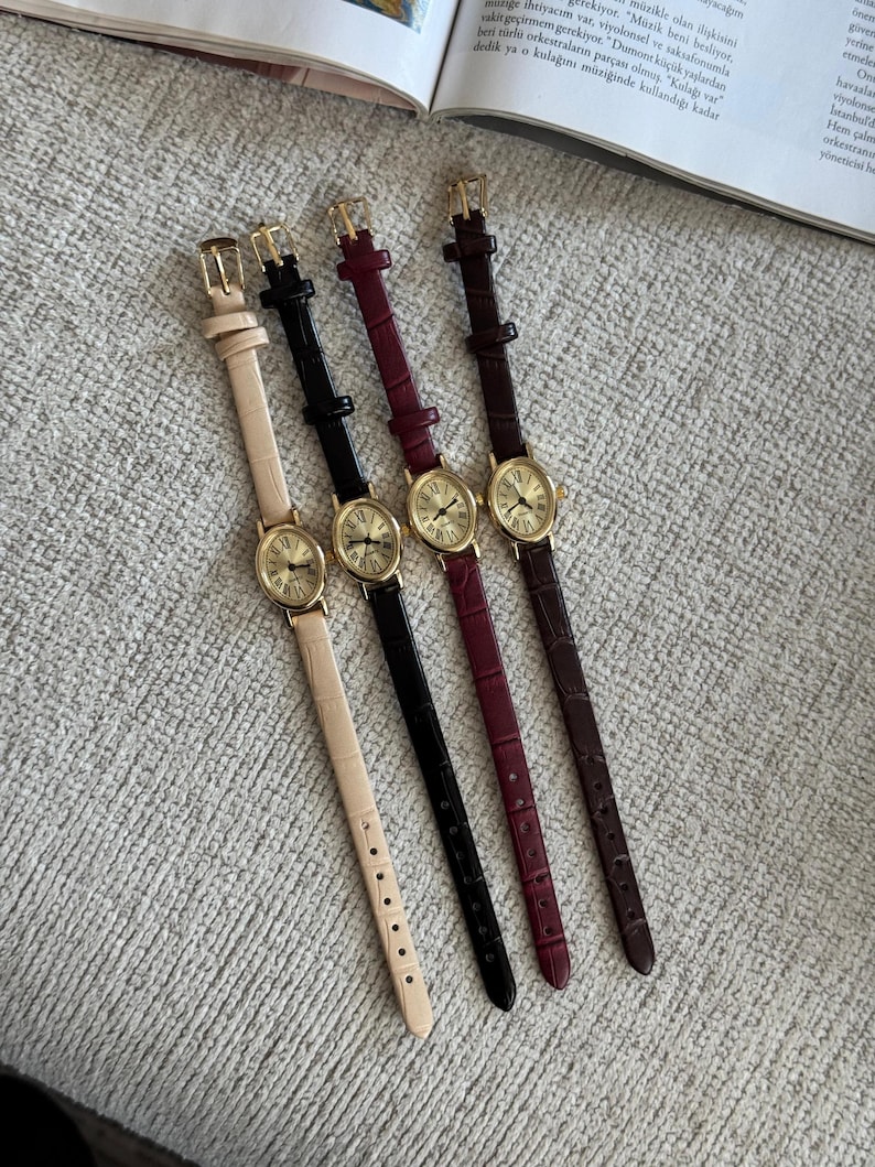 K&ouml;nnte beinhalten: Vier Damenuhren mit goldfarbenen Geh&auml;usen und Lederarmb&auml;ndern in Beige, Schwarz, Burgunderrot und Braun. Jede Uhr hat ein rundes Zifferblatt mit r&ouml;mischen Ziffern und einer kleinen Sekundenscheibe.
