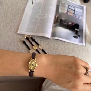 K&ouml;nnte beinhalten: Eine goldfarbene Armbanduhr mit braunem Lederarmband wird am Handgelenk getragen. Die Uhr hat ein ovales Zifferblatt mit r&ouml;mischen Ziffern. Im Hintergrund sind weitere &auml;hnliche Uhren zu sehen. Im Hintergrund ist eine Zeitschrift ge&ouml;ffnet. Das Armband ist ca. 15 cm lang.