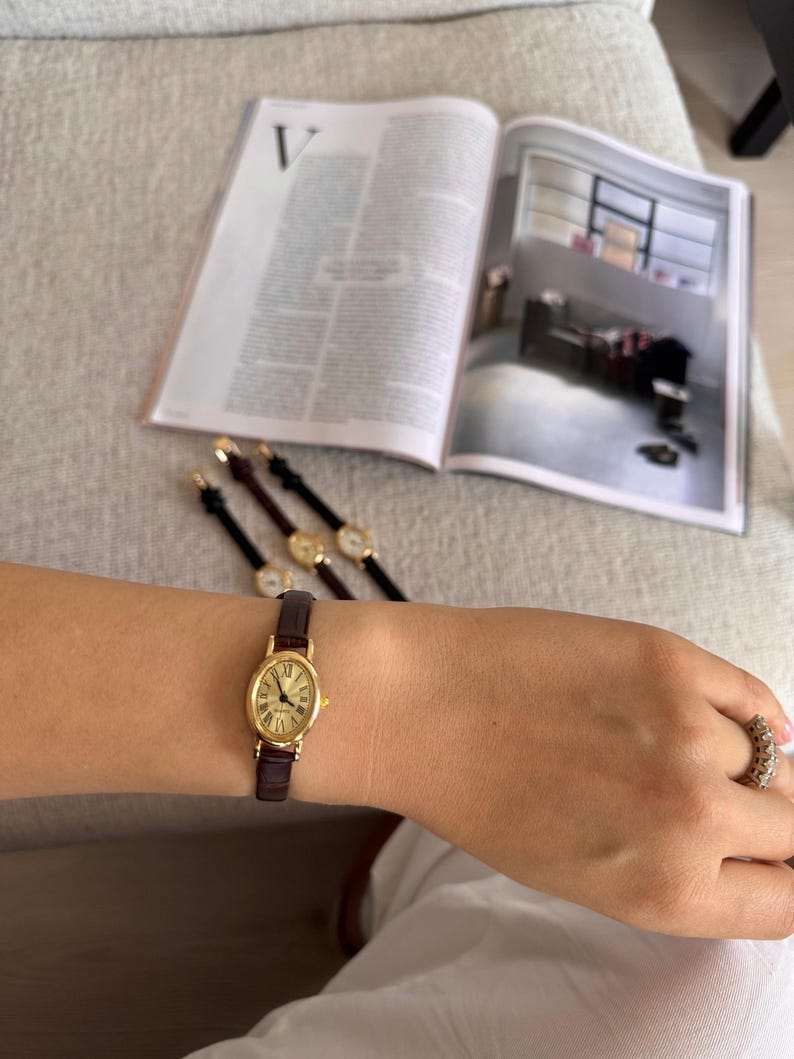 K&ouml;nnte beinhalten: Eine goldfarbene Armbanduhr mit braunem Lederarmband wird am Handgelenk getragen. Die Uhr hat ein ovales Zifferblatt mit r&ouml;mischen Ziffern. Im Hintergrund sind weitere &auml;hnliche Uhren zu sehen. Im Hintergrund ist eine Zeitschrift ge&ouml;ffnet. Das Armband ist ca. 15 cm lang.