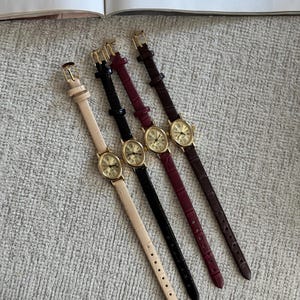 K&ouml;nnte beinhalten: Vier analoge Armbanduhren mit goldfarbenen Geh&auml;usen und Lederarmb&auml;ndern in Beige, Schwarz, Burgunderrot und Braun. Jede Uhr hat ein rundes Zifferblatt mit r&ouml;mischen Ziffern und einen kleinen Sekundenzeiger. Die Uhren liegen auf einer strukturierten, hellgrauen Oberfl&auml;che.