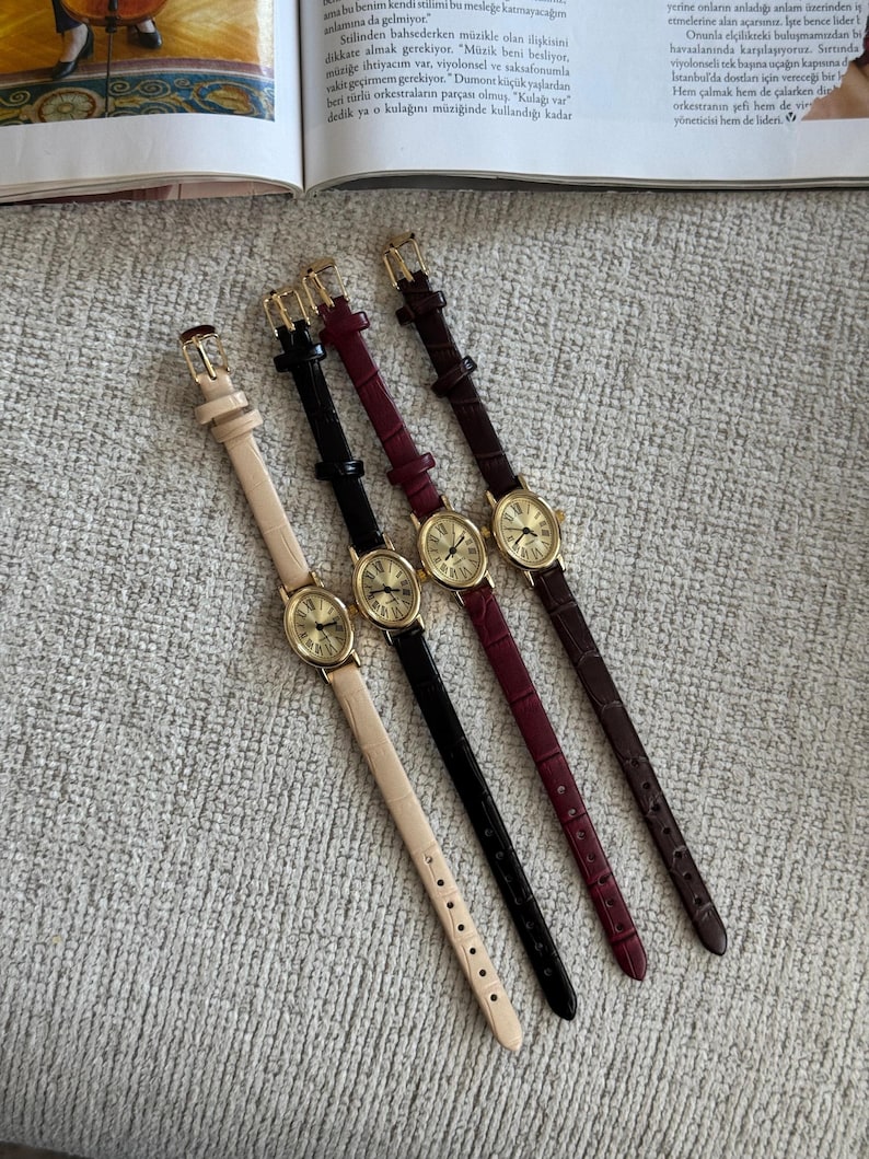 K&ouml;nnte beinhalten: Vier analoge Armbanduhren mit goldfarbenen Geh&auml;usen und Lederarmb&auml;ndern in Beige, Schwarz, Burgunderrot und Braun. Jede Uhr hat ein rundes Zifferblatt mit r&ouml;mischen Ziffern und einen kleinen Sekundenzeiger. Die Uhren liegen auf einer strukturierten, hellgrauen Oberfl&auml;che.