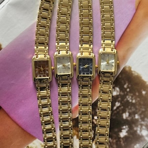 May include: Four gold-coloured rectangular wristwatches with matching link bands are displayed. Each watch has a different coloured face: brown, white, navy blue, and cream. Roman numerals mark the hours. The watches are arranged on a light purple background.