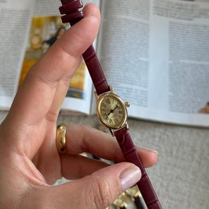 K&ouml;nnte beinhalten: Eine goldfarbene Armbanduhr mit einem weinroten Lederarmband. Das Zifferblatt der Uhr ist rund und mit r&ouml;mischen Ziffern versehen. Das Armband ist etwa 1,3 cm breit. Die Uhr wird in einer Hand gehalten, weitere Uhren sind im Hintergrund zu sehen.