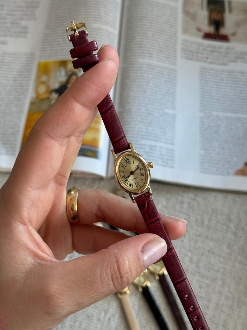 K&ouml;nnte beinhalten: Eine goldfarbene Armbanduhr mit einem weinroten Lederarmband. Das Zifferblatt der Uhr ist rund und mit r&ouml;mischen Ziffern versehen. Das Armband ist etwa 1,3 cm breit. Die Uhr wird in einer Hand gehalten, weitere Uhren sind im Hintergrund zu sehen.
