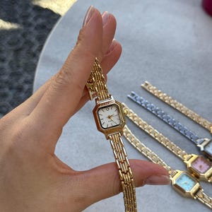 May include: A gold-toned wristwatch with a rectangular face and a linked band. The watch face is white with black hour markers and gold hands. Several other watches with different colored faces and bands are visible in the background.