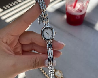 May include: Silver-toned wristwatch with an oval white face and a linked band. The watch has a small, round face with black numerals and hands. Two additional watches are visible in the background, one gold-toned and one silver-toned.