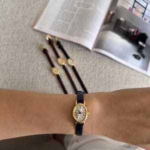 K&ouml;nnte beinhalten: Eine goldfarbene Armbanduhr mit schwarzem Lederarmband wird am Handgelenk getragen. Die Uhr hat ein wei&szlig;es Zifferblatt mit r&ouml;mischen Ziffern. Drei weitere Uhren mit &auml;hnlichem Design sind im Hintergrund zu sehen, zusammen mit einer ge&ouml;ffneten Zeitschrift.