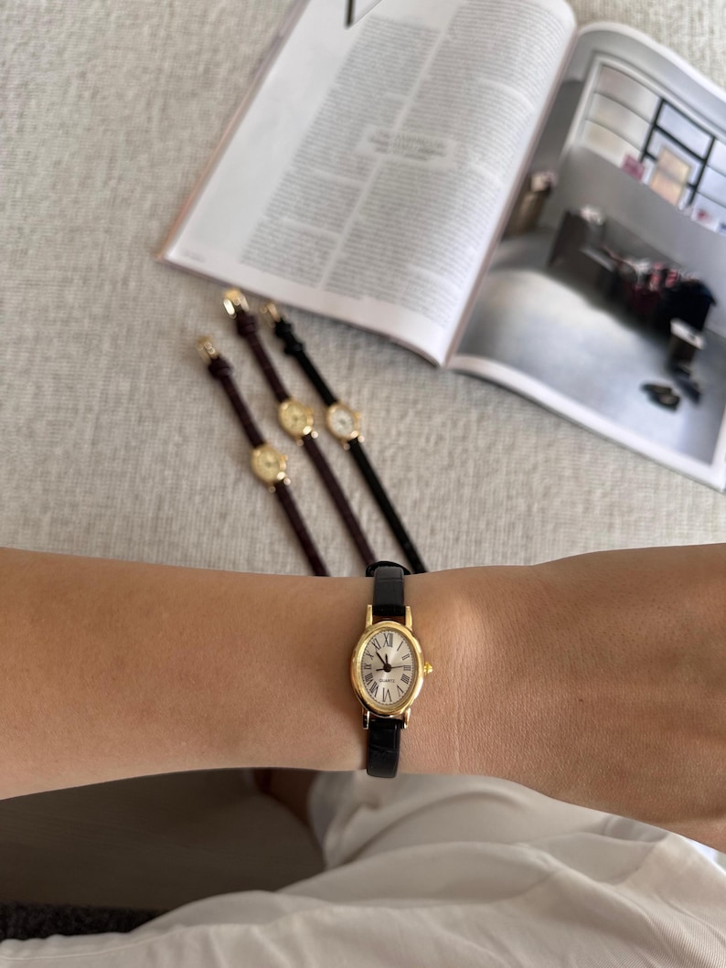 K&ouml;nnte beinhalten: Eine goldfarbene Armbanduhr mit schwarzem Lederarmband wird am Handgelenk getragen. Die Uhr hat ein wei&szlig;es Zifferblatt mit r&ouml;mischen Ziffern. Drei weitere Uhren mit &auml;hnlichem Design sind im Hintergrund zu sehen, zusammen mit einer ge&ouml;ffneten Zeitschrift.