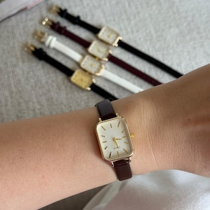 Könnte beinhalten: Eine goldfarbene rechteckige Uhr mit weißem Zifferblatt und braunem Lederarmband wird am Handgelenk getragen. Im Hintergrund sind mehrere ähnliche Uhren mit unterschiedlich farbigen Armbändern zu sehen, darunter schwarz, weiß und bordeaux. Die Uhren haben ein minimalistisches Design.