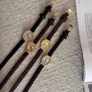 K&ouml;nnte beinhalten: Vier goldfarbene ovale Uhren mit schwarzen und braunen Lederarmb&auml;ndern. Die Zifferbl&auml;tter der Uhren haben r&ouml;mische Ziffern und einen kleinen Sekundenzeiger. Die Armb&auml;nder haben goldfarbene Schnallen. Die Uhren werden auf einer strukturierten Oberfl&auml;che pr&auml;sentiert.