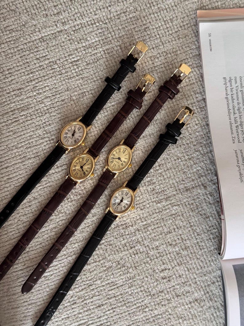 K&ouml;nnte beinhalten: Vier goldfarbene ovale Uhren mit schwarzen und braunen Lederarmb&auml;ndern. Die Zifferbl&auml;tter der Uhren haben r&ouml;mische Ziffern und einen kleinen Sekundenzeiger. Die Armb&auml;nder haben goldfarbene Schnallen. Die Uhren werden auf einer strukturierten Oberfl&auml;che pr&auml;sentiert.