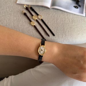 K&ouml;nnte beinhalten: Eine goldfarbene Armbanduhr mit schwarzem Lederarmband wird am Handgelenk getragen. Die Uhr hat ein wei&szlig;es Zifferblatt mit r&ouml;mischen Ziffern. Drei &auml;hnliche Uhren mit unterschiedlich farbigen Armb&auml;ndern sind ebenfalls zu sehen.