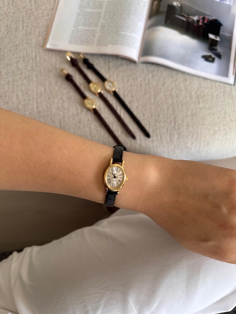 K&ouml;nnte beinhalten: Eine goldfarbene Armbanduhr mit schwarzem Lederarmband wird am Handgelenk getragen. Die Uhr hat ein wei&szlig;es Zifferblatt mit r&ouml;mischen Ziffern. Drei &auml;hnliche Uhren mit unterschiedlich farbigen Armb&auml;ndern sind ebenfalls zu sehen.