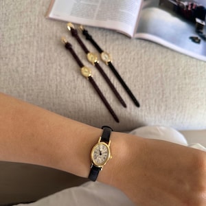 K&ouml;nnte beinhalten: Eine goldfarbene Armbanduhr mit schwarzem Lederarmband wird am Handgelenk getragen. Das Zifferblatt der Uhr ist wei&szlig; mit schwarzen r&ouml;mischen Ziffern. Drei &auml;hnliche Uhren mit verschiedenen Armb&auml;ndern sind ebenfalls sichtbar.