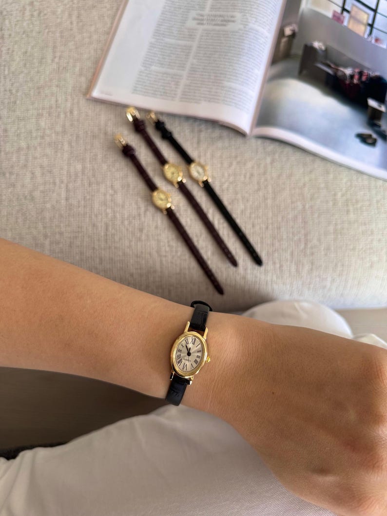 K&ouml;nnte beinhalten: Eine goldfarbene Armbanduhr mit schwarzem Lederarmband wird am Handgelenk getragen. Das Zifferblatt der Uhr ist wei&szlig; mit schwarzen r&ouml;mischen Ziffern. Drei &auml;hnliche Uhren mit verschiedenen Armb&auml;ndern sind ebenfalls sichtbar.