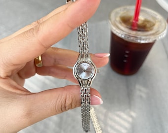 May include: A silver wristwatch with a round, light-colored face and a linked band. The watch is held up, showing the details of the face and band. The watch band is approximately 0.5 inches wide.