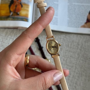 K&ouml;nnte beinhalten: Eine goldfarbene Armbanduhr mit cremefarbenem Lederarmband. Das Zifferblatt der Uhr hat r&ouml;mische Ziffern und goldene Zeiger. Das Armband wird mit einer goldenen Schnalle befestigt. Ein klassisches, elegantes Accessoire.