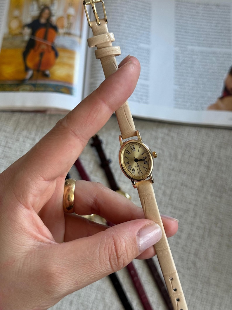 K&ouml;nnte beinhalten: Eine goldfarbene Armbanduhr mit cremefarbenem Lederarmband. Das Zifferblatt der Uhr hat r&ouml;mische Ziffern und goldene Zeiger. Das Armband wird mit einer goldenen Schnalle befestigt. Ein klassisches, elegantes Accessoire.