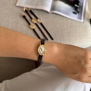 K&ouml;nnte beinhalten: Eine goldfarbene Armbanduhr mit schwarzem Lederarmband wird am Handgelenk getragen. Das Zifferblatt der Uhr ist wei&szlig; mit r&ouml;mischen Ziffern. Im Hintergrund sind drei weitere Uhren mit braunen Armb&auml;ndern zu sehen.