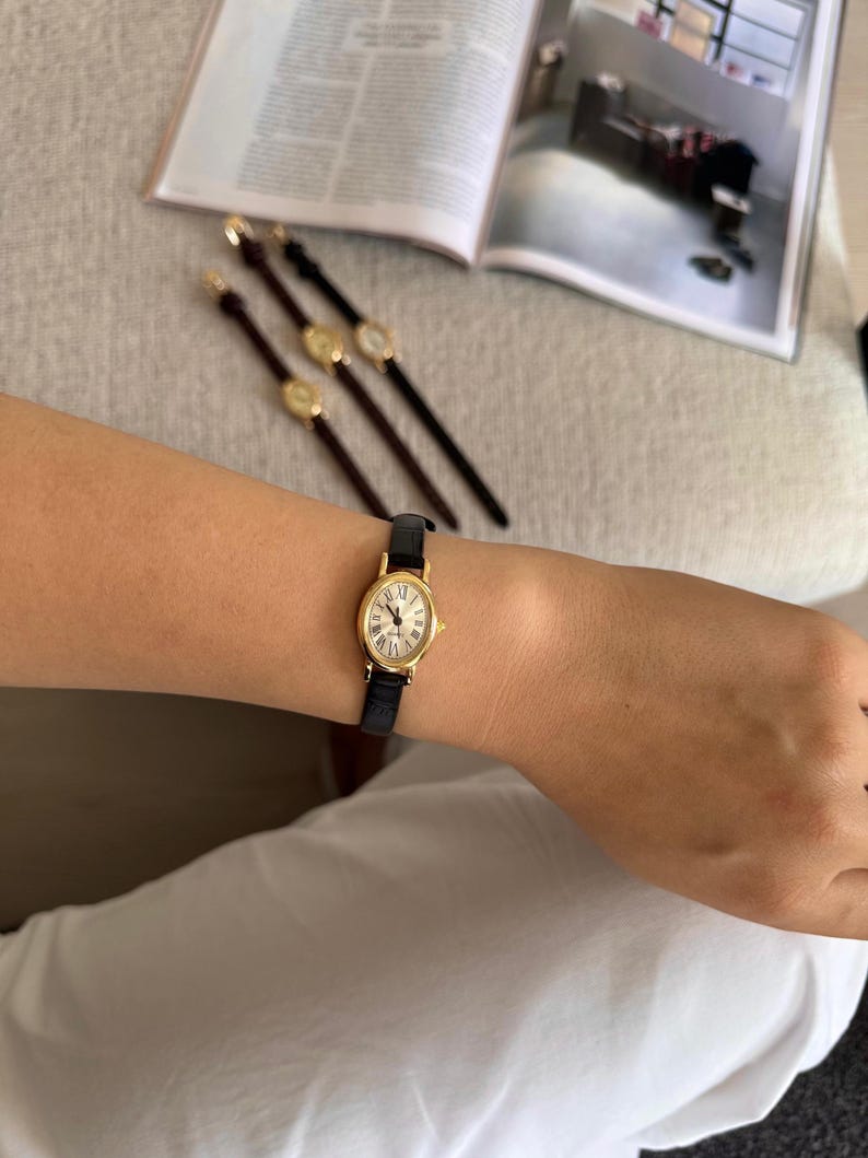 K&ouml;nnte beinhalten: Eine goldfarbene Armbanduhr mit schwarzem Lederarmband wird am Handgelenk getragen. Das Zifferblatt der Uhr ist wei&szlig; mit r&ouml;mischen Ziffern. Im Hintergrund sind drei weitere Uhren mit braunen Armb&auml;ndern zu sehen.