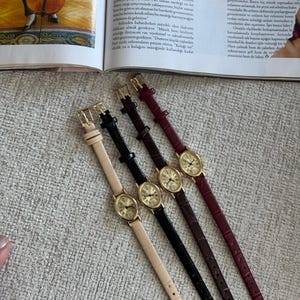 K&ouml;nnte beinhalten: Vier elegante Armbanduhren mit ovalen, goldfarbenen Zifferbl&auml;ttern und Lederarmb&auml;ndern in Beige, Schwarz, Braun und Burgunderrot. Die Uhren sind diagonal angeordnet und pr&auml;sentieren ihr klassisches Design.