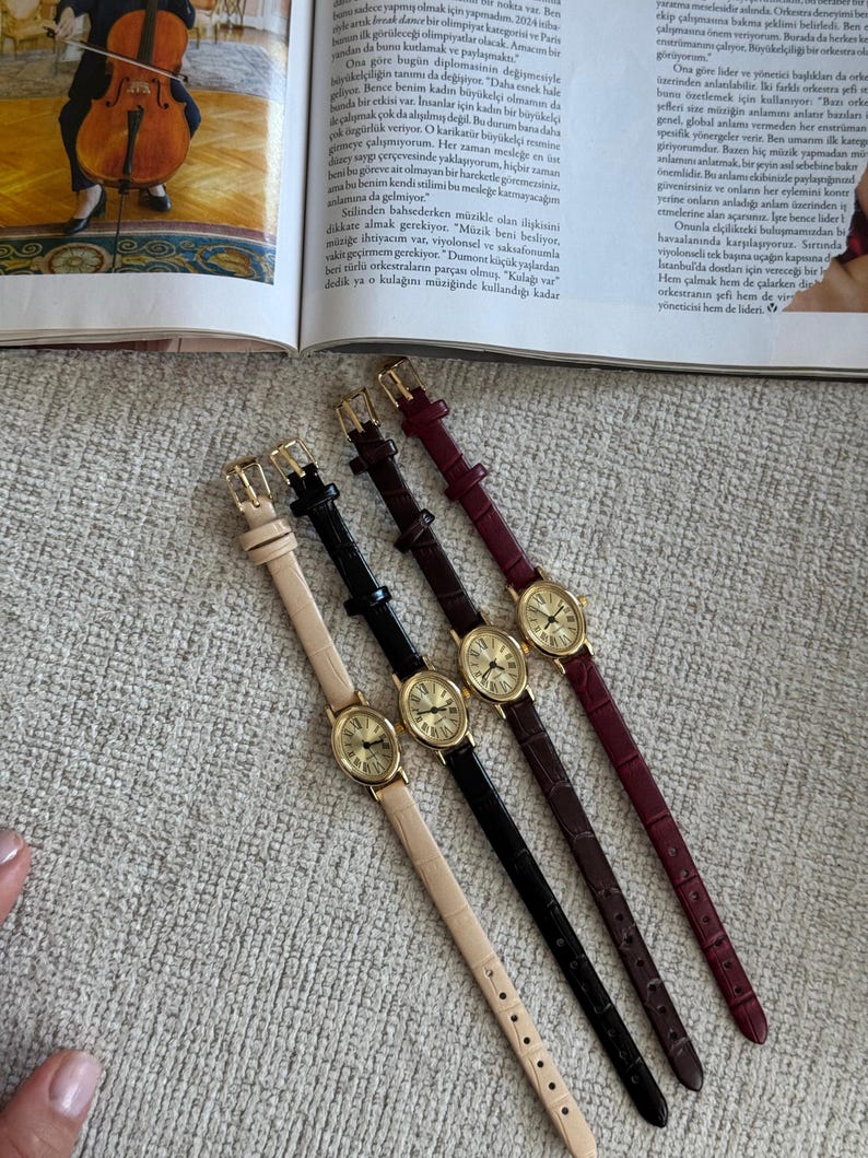 K&ouml;nnte beinhalten: Vier elegante Armbanduhren mit ovalen, goldfarbenen Zifferbl&auml;ttern und Lederarmb&auml;ndern in Beige, Schwarz, Braun und Burgunderrot. Die Uhren sind diagonal angeordnet und pr&auml;sentieren ihr klassisches Design.