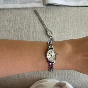 May include: A silver wristwatch with a white face and Roman numeral hour markers is worn on a wrist. The watch has a silver band. Another similar watch is visible in the background.