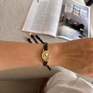 K&ouml;nnte beinhalten: Eine goldfarbene, ovale Armbanduhr mit braunem Lederarmband wird am Arm getragen. Das Zifferblatt der Uhr zeigt r&ouml;mische Ziffern. Die Uhr steht im Mittelpunkt, mit einer Zeitschrift und anderen Armb&auml;ndern im Hintergrund.