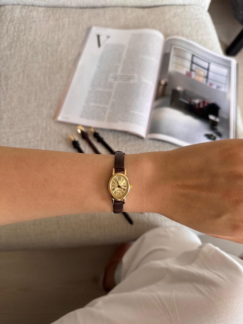 K&ouml;nnte beinhalten: Eine goldfarbene, ovale Armbanduhr mit braunem Lederarmband wird am Arm getragen. Das Zifferblatt der Uhr zeigt r&ouml;mische Ziffern. Die Uhr steht im Mittelpunkt, mit einer Zeitschrift und anderen Armb&auml;ndern im Hintergrund.