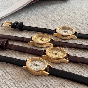 K&ouml;nnte beinhalten: Vier goldfarbene, analoge Armbanduhren mit Lederarmb&auml;ndern. Zwei Uhren haben schwarze Armb&auml;nder und zwei haben braune Armb&auml;nder. Die Zifferbl&auml;tter sind entweder goldfarben oder wei&szlig; mit schwarzen r&ouml;mischen Ziffern und Zeigern. Die Uhren liegen auf einer strukturierten, hellen Oberfl&auml;che.