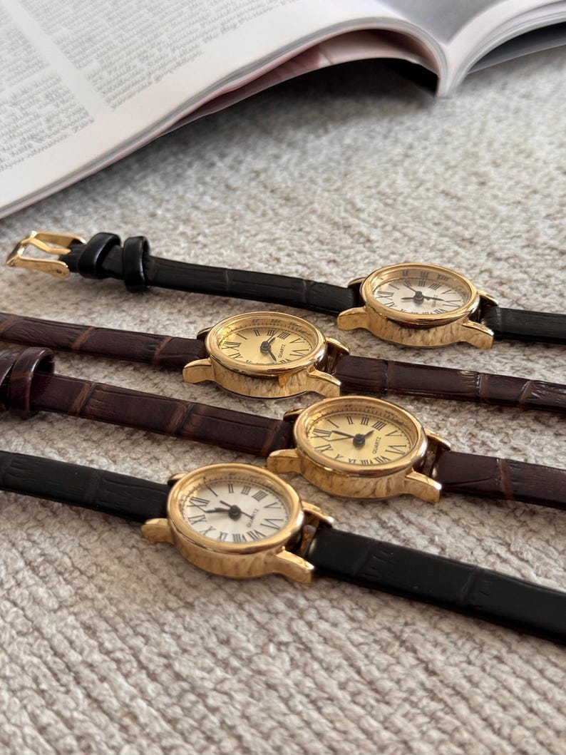 K&ouml;nnte beinhalten: Vier goldfarbene, analoge Armbanduhren mit Lederarmb&auml;ndern. Zwei Uhren haben schwarze Armb&auml;nder und zwei haben braune Armb&auml;nder. Die Zifferbl&auml;tter sind entweder goldfarben oder wei&szlig; mit schwarzen r&ouml;mischen Ziffern und Zeigern. Die Uhren liegen auf einer strukturierten, hellen Oberfl&auml;che.