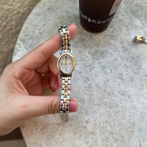 May include: A gold and silver-toned wristwatch with a white face and gold hands. The watch has a rectangular link band. The watch is held in a hand.