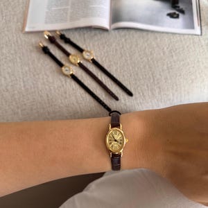 K&ouml;nnte beinhalten: Eine ovale, goldfarbene Armbanduhr mit braunem Lederarmband wird am Handgelenk getragen. Drei &auml;hnliche Uhren mit unterschiedlich farbigen Armb&auml;ndern sind auf einer hellgrauen Oberfl&auml;che angeordnet. Das Zifferblatt der Uhr hat r&ouml;mische Ziffern.