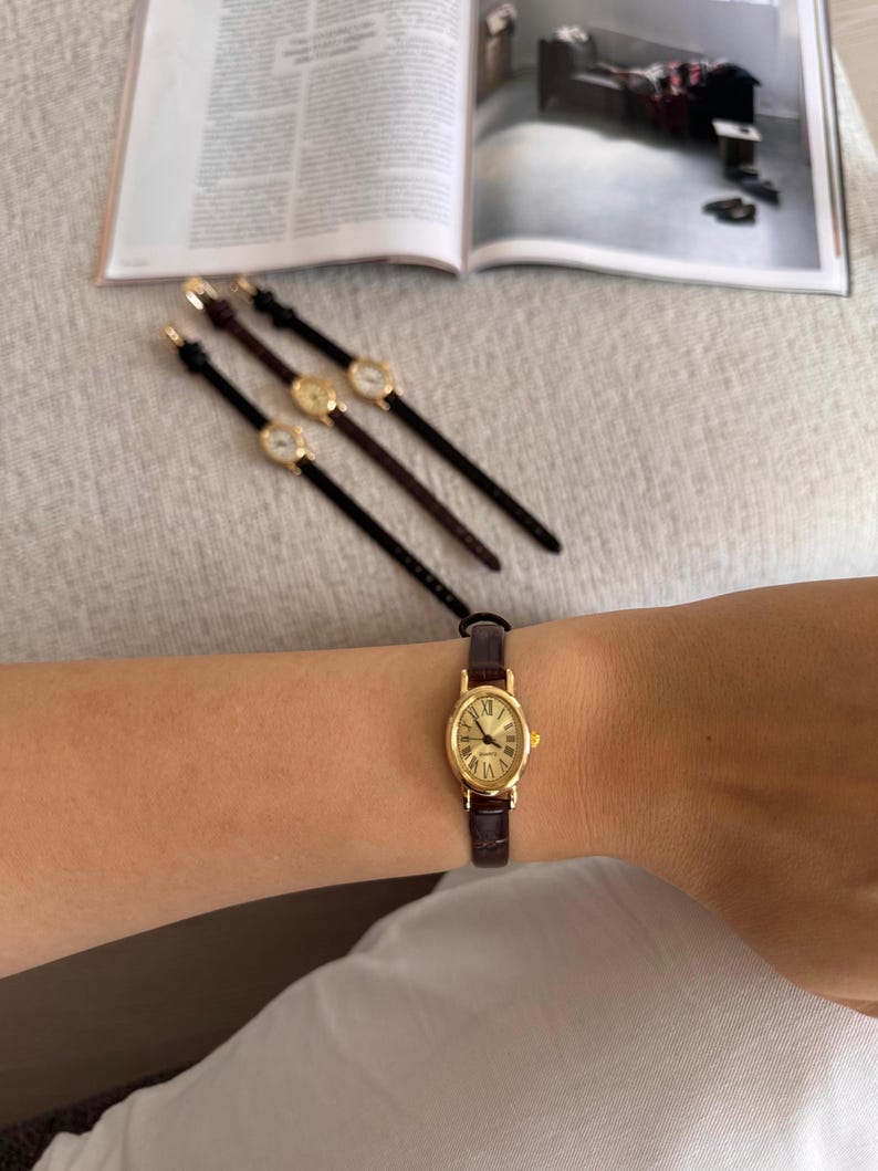 K&ouml;nnte beinhalten: Eine ovale, goldfarbene Armbanduhr mit braunem Lederarmband wird am Handgelenk getragen. Drei &auml;hnliche Uhren mit unterschiedlich farbigen Armb&auml;ndern sind auf einer hellgrauen Oberfl&auml;che angeordnet. Das Zifferblatt der Uhr hat r&ouml;mische Ziffern.