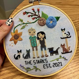 Könnte beinhalten: Ein Kreuzstich-Stickrahmen mit einem Paar, Haustieren und floralen Akzenten. Das Design umfasst eine Sonnenblume, eine blaue Blume und den Text "THE STARKS EST. 2021". Der Rahmen befindet sich in einem Holzrahmen.
