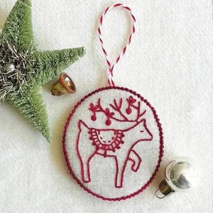May include: Embroidered Christmas ornament featuring a red reindeer design on a circular, off-white fabric. The ornament has a red stitched border and a red and white twisted cord for hanging. A green star and a small bell are also visible.