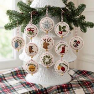 May include: A collection of round, embroidered Christmas ornaments with various festive designs, including a snowflake, holly, and a reindeer. Each ornament is trimmed with red and white thread and has a small gold bell. They are displayed on a white Christmas tree.