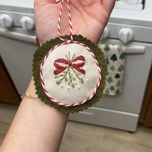 May include: Embroidered Christmas ornament featuring a red bow and mistletoe design on a cream-colored fabric. The ornament is trimmed with red and white cord and a green felt backing. A festive holiday decoration.