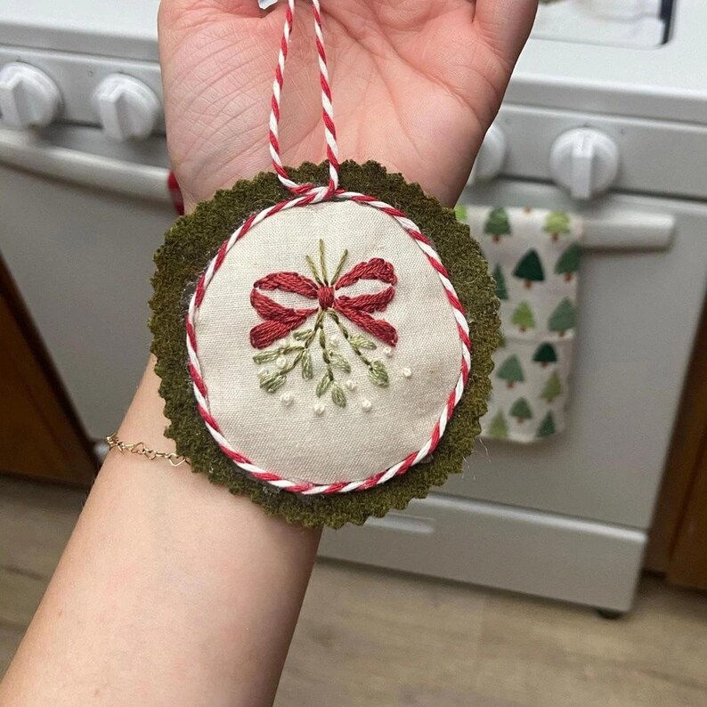 May include: Embroidered Christmas ornament featuring a red bow and mistletoe design on a cream-colored fabric. The ornament is trimmed with red and white cord and a green felt backing. A festive holiday decoration.
