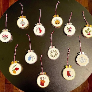 May include: A collection of embroidered Christmas ornaments. Each ornament is round, featuring festive designs like a Christmas tree, snowflake, and holly. They have a red and white striped hanging cord and are set against a dark background.