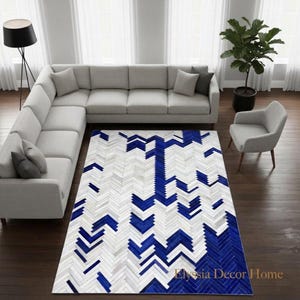 May include: A rectangular area rug featuring a geometric herringbone pattern in alternating shades of white, light grey, and vibrant blue. The rug is composed of small, rectangular segments arranged in a repeating chevron-like layout.