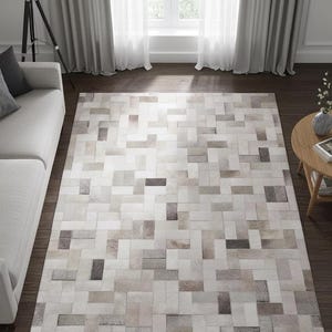 May include: A rectangular area rug with a patchwork design. The rug features a pattern of rectangular shapes in shades of white, gray, and brown. The rug is placed on a dark wood floor, and the room has a neutral color scheme.