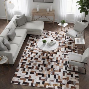 Könnte beinhalten: Innenaufnahme eines Wohnzimmers mit einem hellgrauen Ecksofa, Sesseln und einem runden Marmor-Couchtisch. Ein Patchwork-Teppich in Braun-, Schwarz- und Weißtönen bedeckt den Boden. Der Raum ist hell und natürlich beleuchtet.