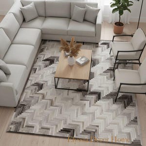 Peut inclure: Aménagement de salon moderne avec un canapé d'angle gris clair et des coussins assortis, deux fauteuils et une table basse en bois. Un tapis à motifs avec un motif en chevrons dans les tons gris et marron recouvre le sol. Une plante en pot et un lampadaire complètent la scène.