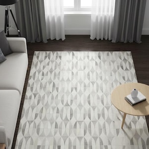 May include: A rectangular area rug with a geometric pattern in shades of gray and white. The rug features an oval design and is placed on a dark wood floor. The room includes a light-colored sofa, a round wooden coffee table, and gray curtains.