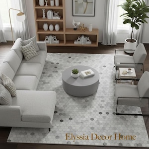 May include: A modern living room with a light gray sectional sofa, two armchairs, and a round coffee table. A hexagonal patterned rug covers the floor. A wooden bookshelf and artwork are in the background. The text "Elyssia Decor Home" is on the rug.