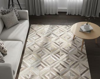 May include: Geometric patterned area rug in shades of beige, gray, and brown. The rug features a repeating pattern of angled lines and shapes. A light gray sofa with decorative pillows is to the left. A round wooden coffee table is to the right.