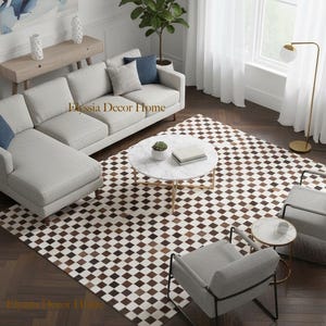 May include: An interior shot of a modern living room with a light gray sectional sofa, two gray armchairs, and a marble-topped coffee table. A patterned rug with brown and white squares covers the floor. A gold floor lamp and a painting are also visible.